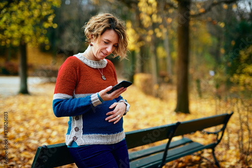 Pregnant blond woman in a park looking at her cell phone and smiling while holding her belly. The park is autumn yellow.