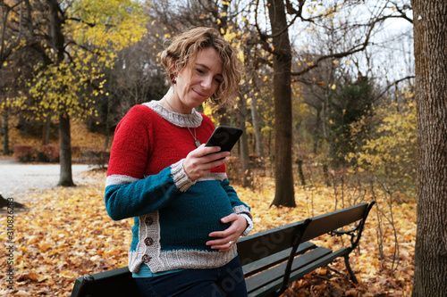 Pregnant blond woman in a park looking at her cell phone and smiling while holding her belly. The park is autumn yellow.