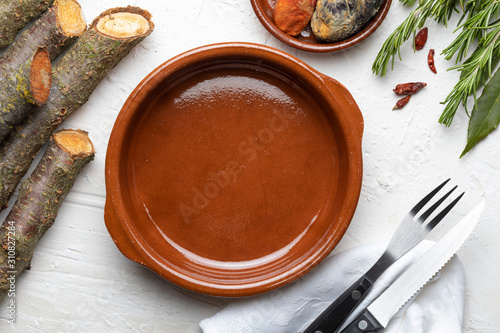 Empty clay pot. On white wooden background. Of rustic aspect it includes spices (rosemary, laurel, chilli peppers) and cutlery. Concept to include your food and your text.