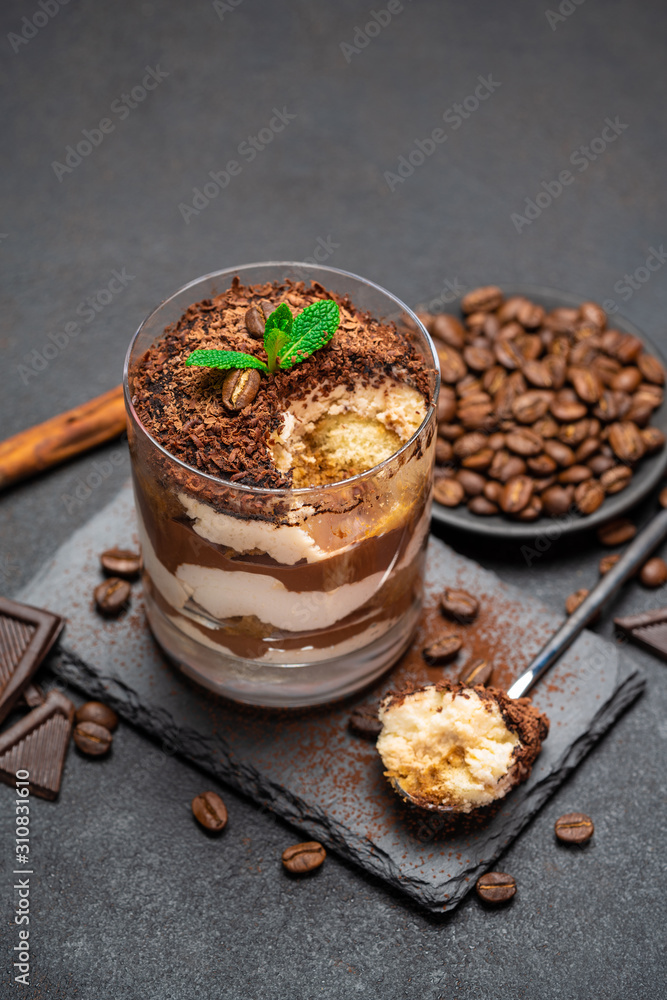 Classic tiramisu dessert in a glass cup and pieces of chocolate on stone cutting board on dark concrete background Classic tiramisu dessert in a glass cup and pieces of chocolate on stone cutting board on dark concrete background