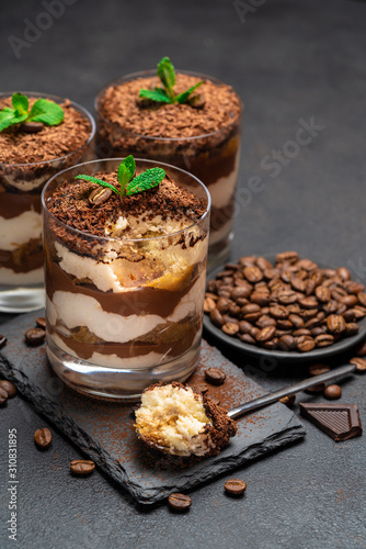 Classic tiramisu dessert in a glass cup and pieces of chocolate on stone cutting board on dark concrete background