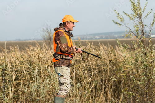 A man with a gun in his hands and an orange vest on a pheasant hunt in a wooded area in cloudy weather. Hunter with dogs in search of game. © Mountains Hunter