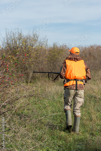 A man with a gun in his hands and an orange vest on a pheasant hunt in a wooded area in cloudy weather. Hunter with dogs in search of game.