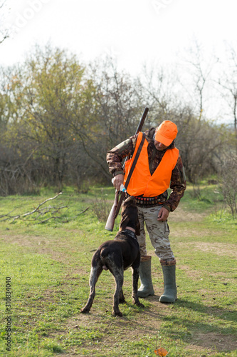 A man with a gun in his hands and an orange vest on a pheasant hunt in a wooded area in cloudy weather. Hunter with dogs in search of game.