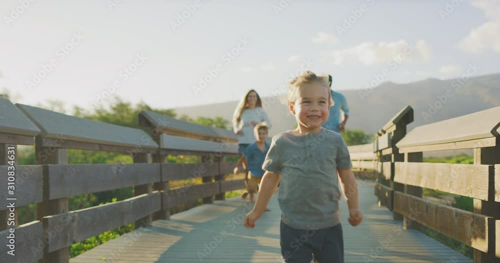 Happy smiling toddler boy runs joyfully in front of his brother and parents on a coastal boardwalk at sunset, weekend family fun, happy family running on a boardwalk Happy smiling toddler boy runs joyfully in front of his brother and parents on a coastal boardwalk at sunset, weekend family fun, happy family running on a boardwalk