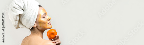 Beauty portrait of woman in white towel on head with gold nourishing mask on face. Skincare cleansing eco organic cosmetic spa relax concept. A girl stands with her back holding an orange mandarin.