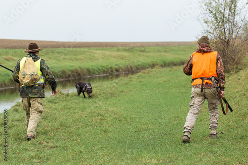 A mans with a gun in his hands and an orange vest on a pheasant hunt in a wooded area in cloudy weather. Hunters with dogs in search of game.