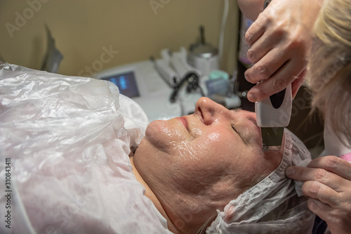 A cosmetic procedure with ultrasonic cleaning of the skin of the face is performed for an aged woman. Instrumental effect on the surface of the skin. A series of sequential operations.