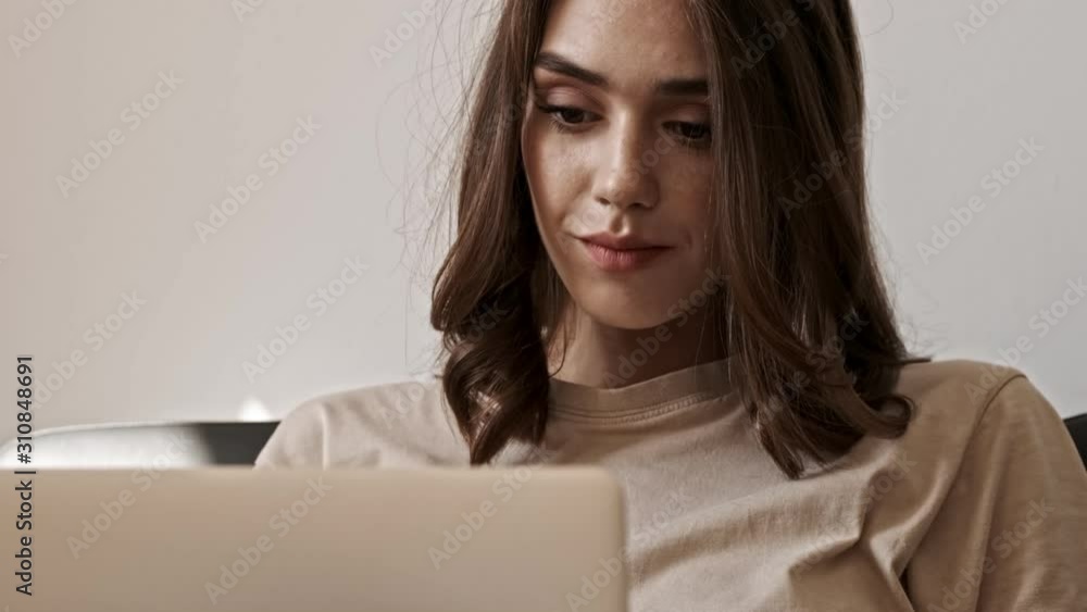 Smiling concentrated brunette woman using laptop computer and looking away after that while sitting on sofa indoors Smiling concentrated brunette woman using laptop computer and looking away after that while sitting on sofa indoors