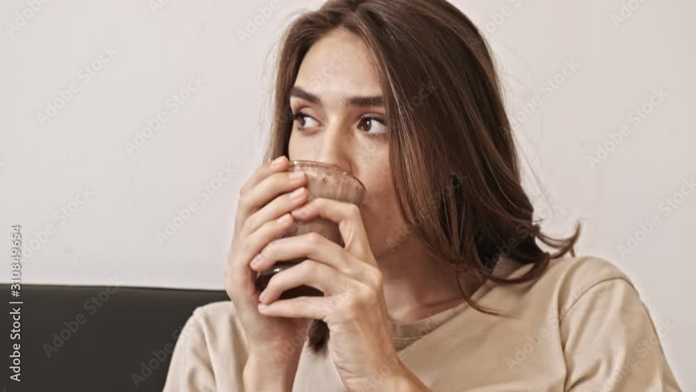 Close up view of smiling pretty brunette woman drinking cocoa and looking away while sitting on sofa indoors Close up view of smiling pretty brunette woman drinking cocoa and looking away while sitting on sofa indoors