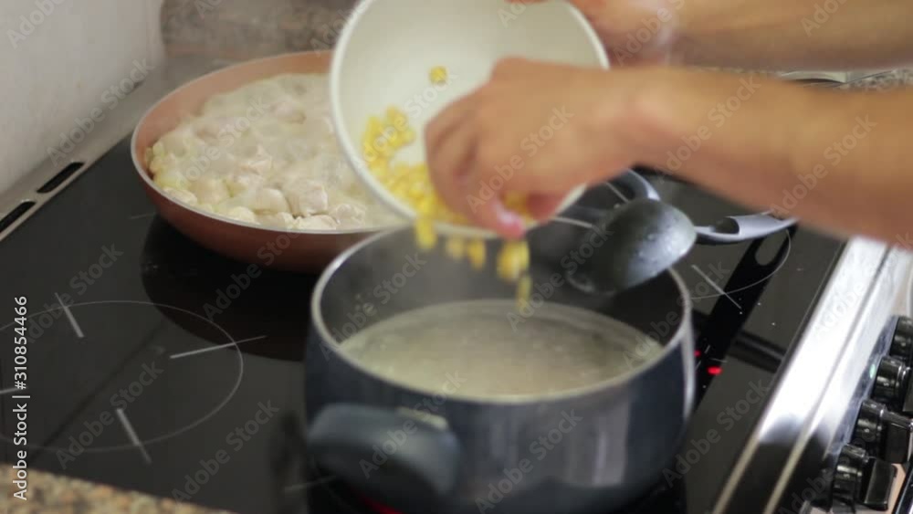 Pouring Corn into Pot of Boiling Water and Stirring With Chicken in Frying Pan in Background Pouring Corn into Pot of Boiling Water and Stirring With Chicken in Frying Pan in Background