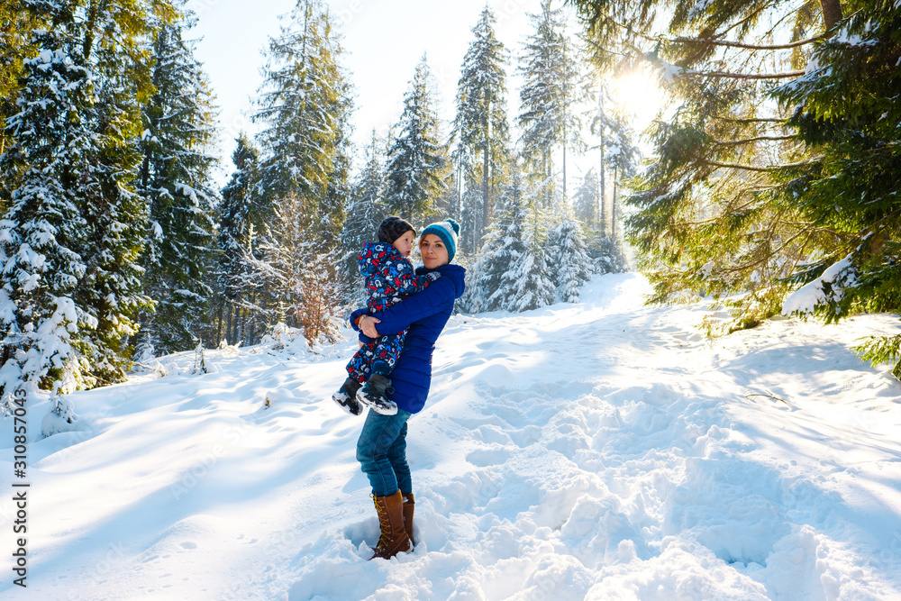 Happy mother and child boy having fun, walking in snowy winter forest. Family spend time together outdoors. Mom with little kid playing with snow and smiling in park. Christmas holiday with children.