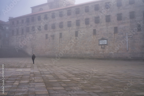 A person walking alone through the streets of an ancient city next to a Catholic monastery on an autumn day with thick fog in the city. Plaza da Quintana, Santiago de Compostel. Galicia, Spain.