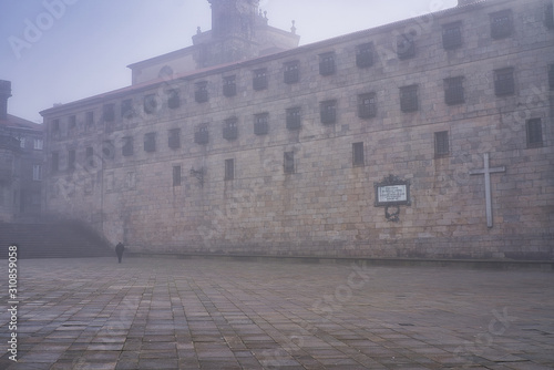 A person walking alone through the streets of an ancient city next to a Catholic monastery on an autumn day with thick fog in the city. Plaza da Quintana, Santiago de Compostel. Galicia, Spain.