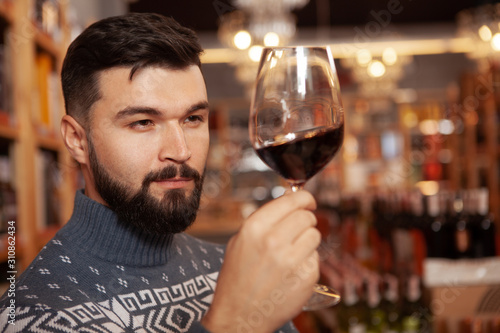 Close up of a handsome bearded man examining red wine in his glass, tasting drinks at the winery