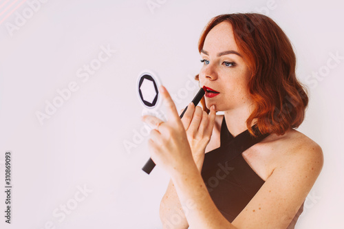 Attractive red-haired girl doing lips makeup. Studio portrait of beautiful woman in elegant black dress, using lipstick and mirror.