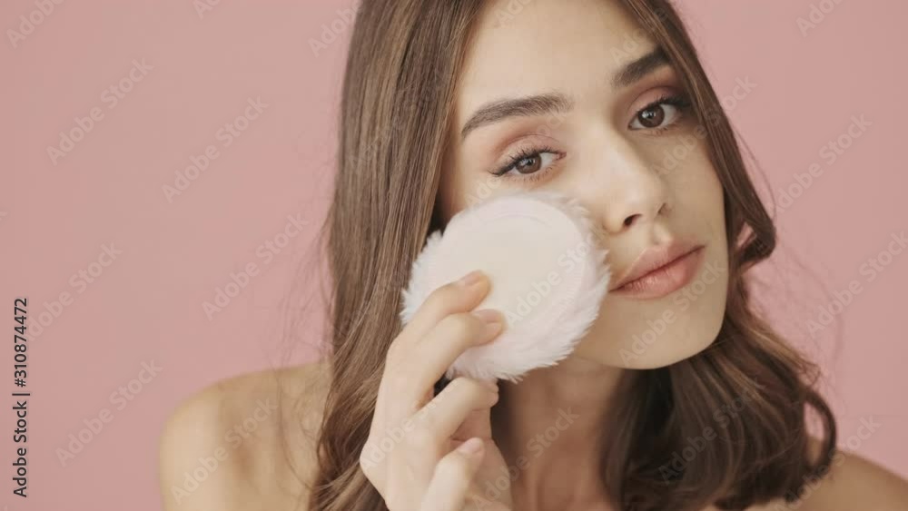 A stylish half-naked woman is applying makeup with powder puff isolated over pink background A stylish half-naked woman is applying makeup with powder puff isolated over pink background