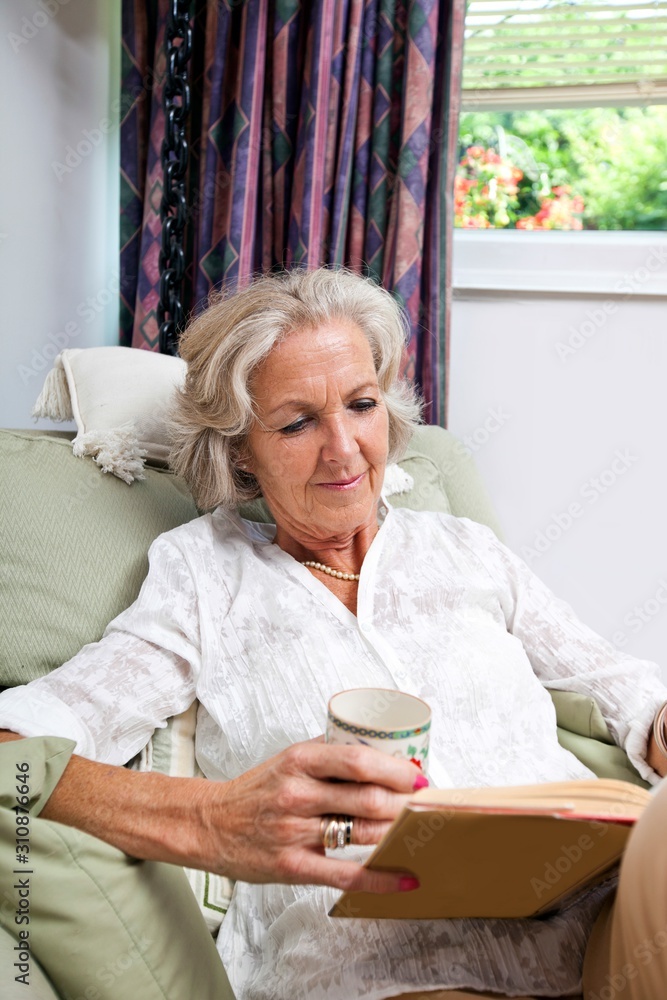 Senior woman with coffee cup reading book while relaxing on armchair at home Senior woman with coffee cup reading book while relaxing on armchair at home