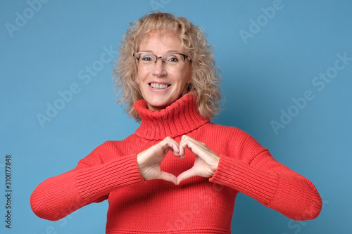 Smiling mature blonde woman girl inred sweater posing showing shape heart with hands. Love emotion or healthy lifestyle concept. photo