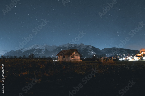 Wooden Hut With Snow Mountains In The Background At Night Time In The Middle On The Frame.