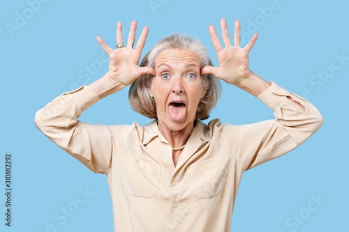 Fotografie Portrait of senior woman making funny faces with hands on head against blue back