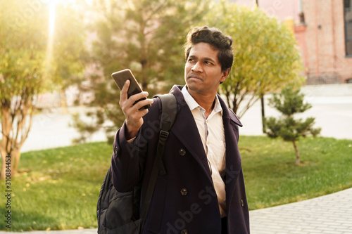 A man in a dark blue jacket and black backpack is standing in city with a phone in his hands looking for navigation. Solo overseas traveler, sightseeing. Taxi booking by mobile app