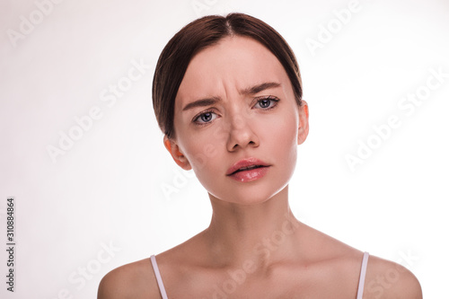 Worried young woamn look straight on camera. Show wrinkles between eyebrowes. Brunette look straight. Slim woman with naked shoulders. Anti-aging. Isolated over light background.