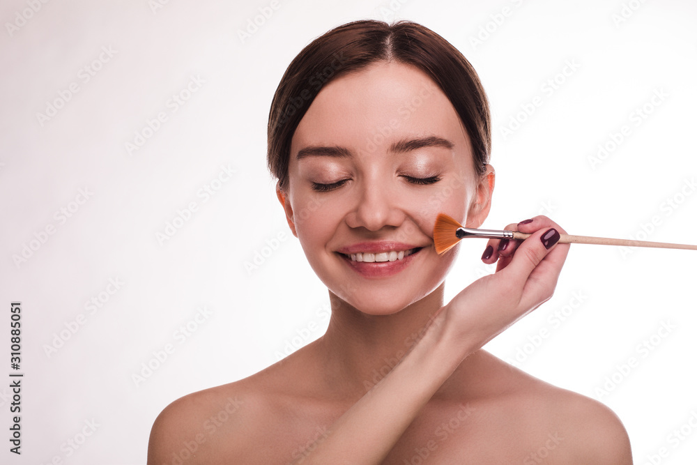 Cheerful attractive young woman with clear skin and brown hair smile with closed eyes. Hand hold make up brush and put on blush or powder. Cosmetics and skincare. Isolated on white background. Cheerful attractive young woman with clear skin and brown hair smile with closed eyes. Hand hold make up brush and put on blush or powder. Cosmetics and skincare. Isolated on white background.
