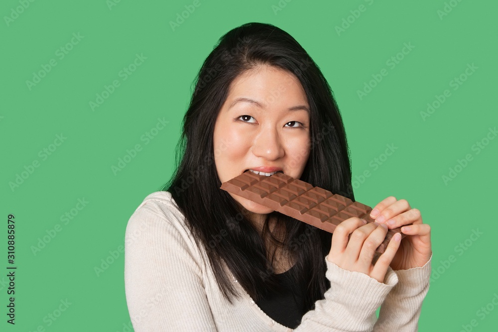 Cute young woman eating a large chocolate bar over green background Cute young woman eating a large chocolate bar over green background