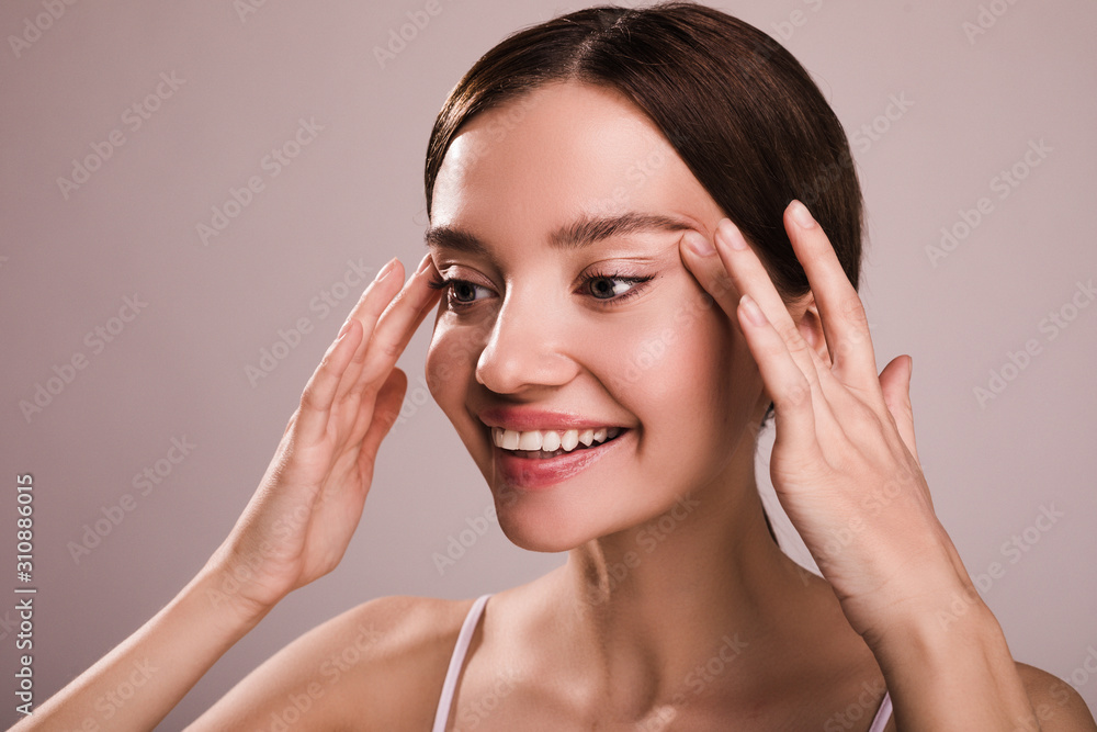 Fototapeta premium Happy positive young woman look to left and smile. Hold hands aside of head. Cheerful positive model touching skin close to eyes. Isolated over beige background. Wrinkles concept. Anti-age