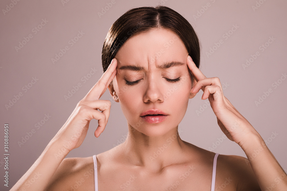 Disturbed worried young woman keep eyes closed and fingers close to head. Has headache or pain. Suffer from discomfort. Young model without pain relief. Isolated over beige background. Disturbed worried young woman keep eyes closed and fingers close to head. Has headache or pain. Suffer from discomfort. Young model without pain relief. Isolated over beige background.