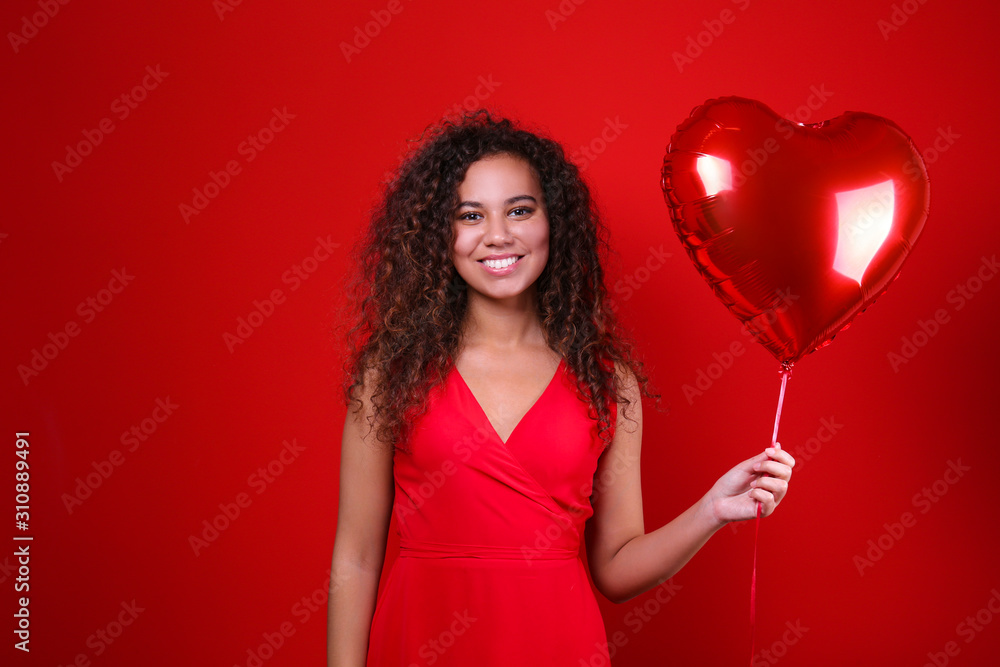 Studio portrait of young woman with dark skin and long curly hair wearing sexy dress over the festive red wall with heart shaped balloon. Close up, isolated background, copy space. Studio portrait of young woman with dark skin and long curly hair wearing sexy dress over the festive red wall with heart shaped balloon. Close up, isolated background, copy space.