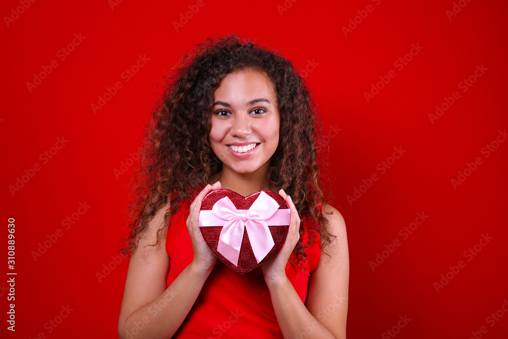 Studio portrait of young woman with dark skin and long curly hair wearing sexy dress over the festive red wall holding heart shaped candy box. Close up, isolated background, copy space. Studio portrait of young woman with dark skin and long curly hair wearing sexy dress over the festive red wall holding heart shaped candy box. Close up, isolated background, copy space.