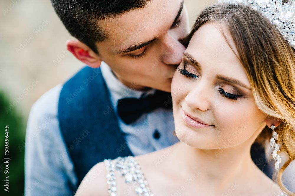 Fototapeta premium Stylish happy bride, woman with crown and groom, man. Newlyweds are hugging and kiss. Close up. On the background of nature. Weddings, engagement, marriage proposal, dating.