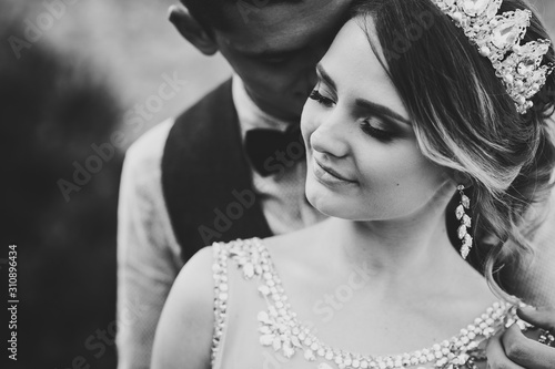 Stylish happy bride, woman with crown and groom, man. Newlyweds are hugging and kiss. Close up. On the background of nature. Black and white photo