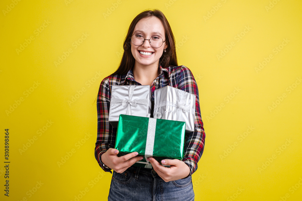 Happy beautiful girl in plaid shirt holding gift boxes enjoying gifts standing on yellow background Happy beautiful girl in plaid shirt holding gift boxes enjoying gifts standing on yellow background