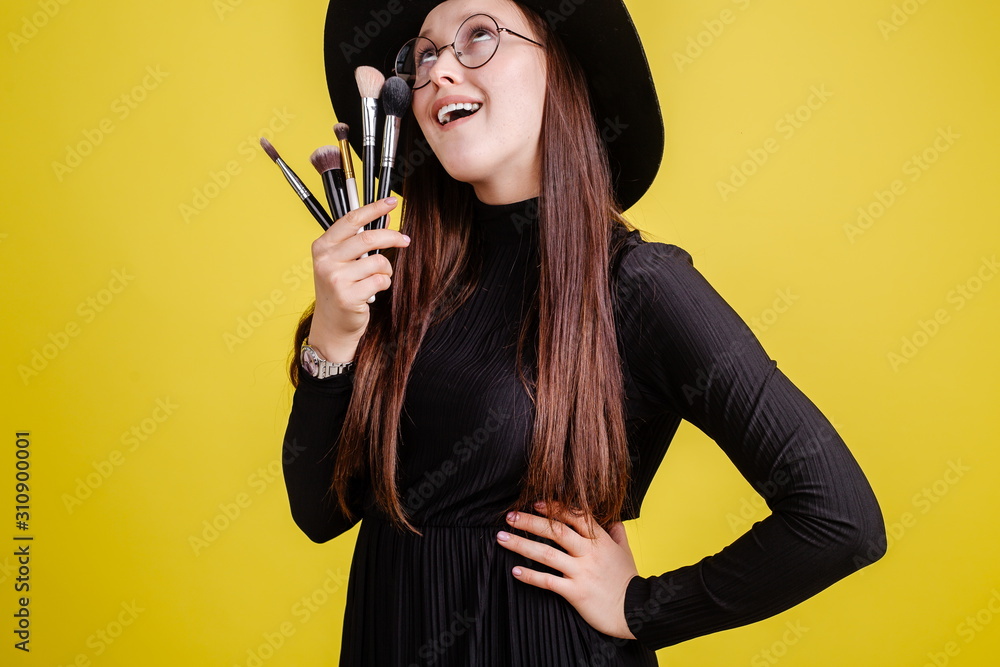 Beautiful girl in a black hat with glasses for vision, holding makeup brushes in front of her standing on a yellow background Beautiful girl in a black hat with glasses for vision, holding makeup brushes in front of her standing on a yellow background