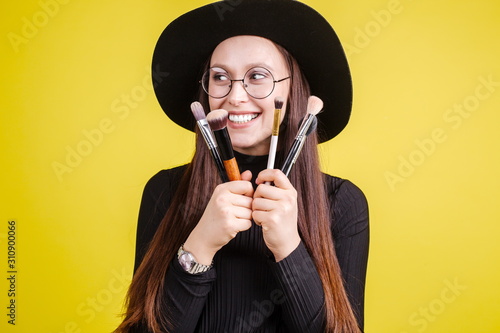 Beautiful girl in a black hat with glasses for vision, holding makeup brushes in front of her