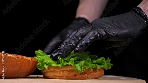 Craft burger is cooking on black background in black food gloves. Consist: sauce, lettuce, tomato, red onion, pickle, cheese, bacon, air bun and marbled meat beef. Not made ideal. Looks real, loving