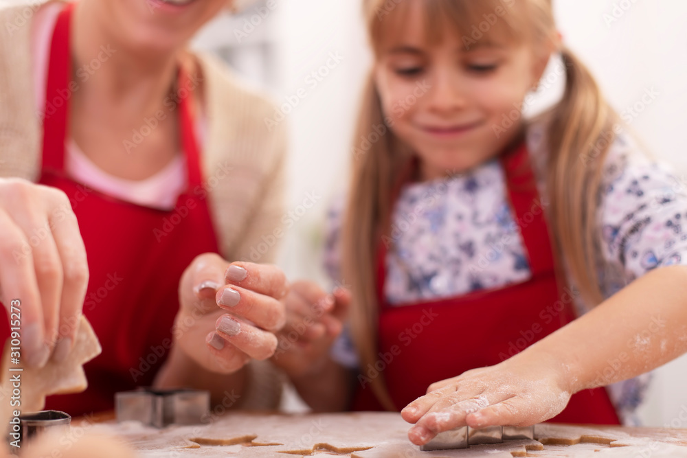 Fototapeta premium Cute little girl cutting gingerbread cookies from the stretched dough - her mother assisting and helping