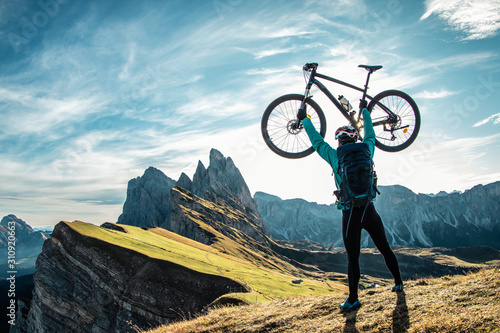 Obraz na plátně Young man raising mountain bike to the sky on Seceda mountain peak at sunrise
