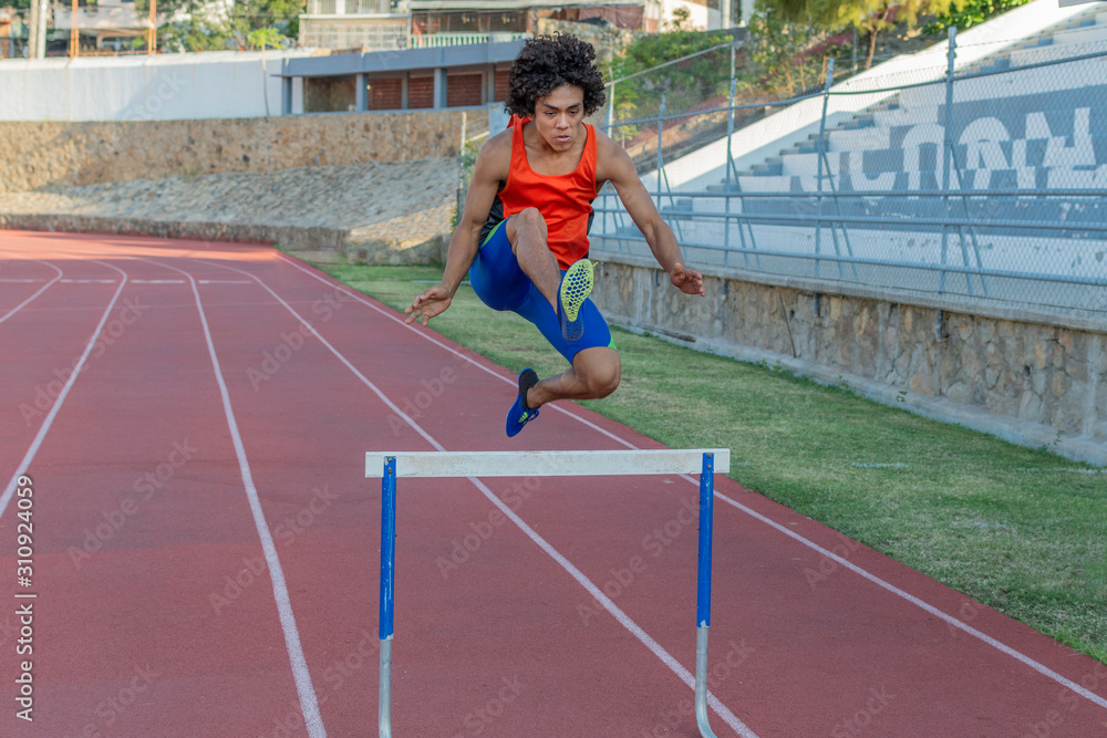 Latin fit young man with afro style hair in orange tank top at the running track jumping the hurdless fence on a sunny day.