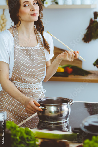 Young brunette woman cooking soup in kitchen. Housewife holding wooden spoon in her hand. Food and health concept