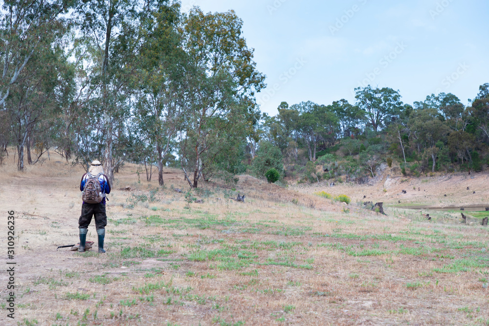 A fisherman walking through dry bushland towards a fresh water reservoir to locate a suitable fishing spot A fisherman walking through dry bushland towards a fresh water reservoir to locate a suitable fishing spot