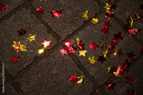 Shiny star shaped tinsel decorations atop rough background of grey stone tiled floor surface. Abstract backdrop with glowing red and golden foil stars. Birthday party theme. New Year celebration decor
