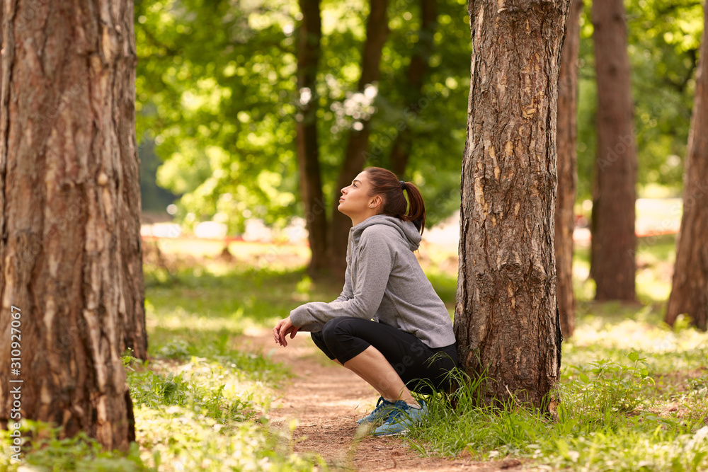 Side view of beautiful caucasian brunette with ponytail and in sportswear crouching in woods and resting after running. Side view of beautiful caucasian brunette with ponytail and in sportswear crouching in woods and resting after running.