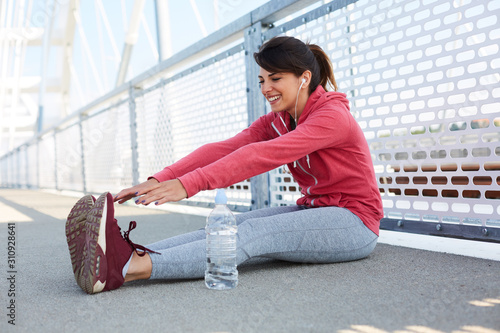 Beautiful smiling caucasian brunette in sportswear sitting on the bridge, listening music and stretching. Outdoor fitness concept.