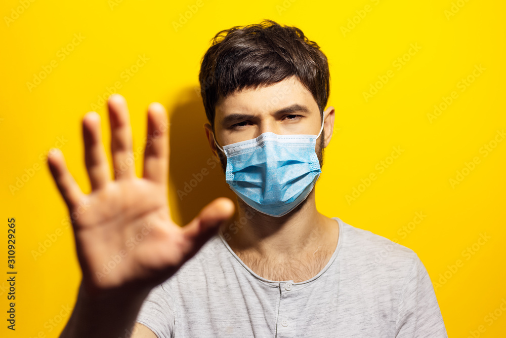 Portrait of young ill man wearing medical flu mask, shows gesture STOP with hand on background of yellow. Portrait of young ill man wearing medical flu mask, shows gesture STOP with hand on background of yellow.