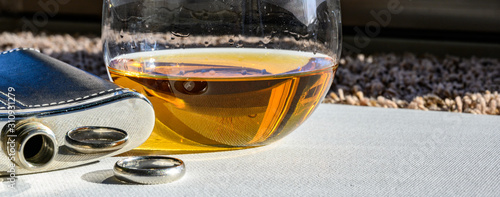 Black and silver whiskey flask next to a glass of amber colored liquid and a wedding ring