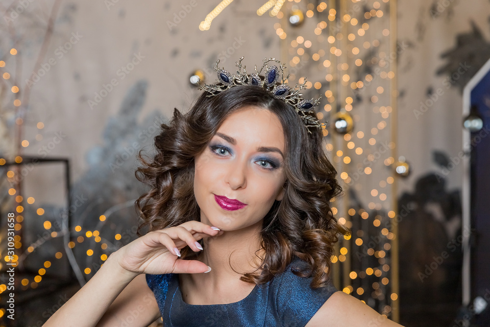 Fototapeta premium Beautiful brunette girl with a golden crown. Beauty face.Fashion Portrait of Beautiful Girl in exquisite jewelry. Beauty sexy Portrait. model with red lips in a crown and earrings is looking at the